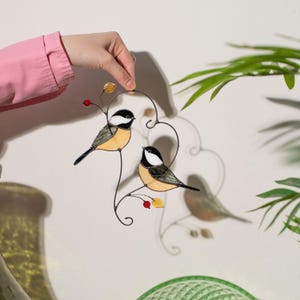 Stained glass bird suncatcher held by hand casting shadow on wall, handmade chickadee glass decor with berries and leaves, artistic window hanging.