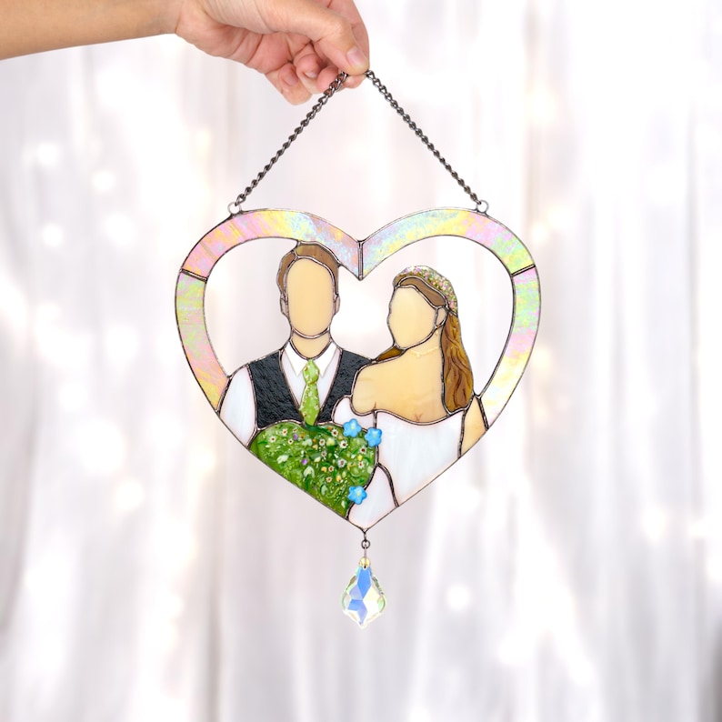 Hand holding a heart-shaped stained glass portrait of a bride and groom with a crystal drop, displayed against white curtains.