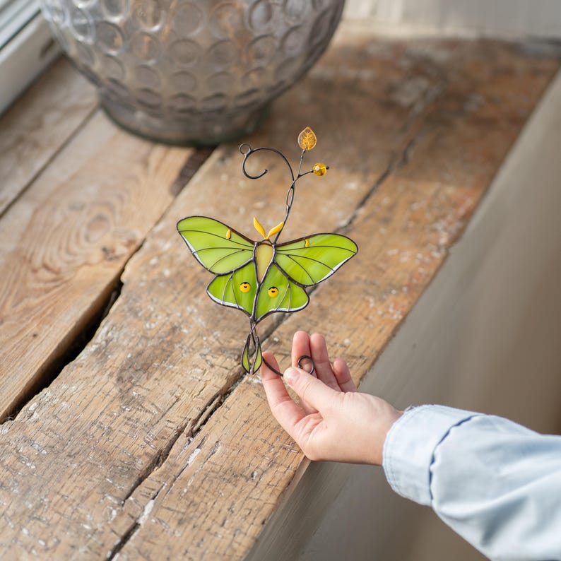 A Luna Moth stained glass suncatcher resting on a rustic wooden table. A hand is reaching out to pick it up, emphasizing its craftsmanship as a high-quality garden decoration.