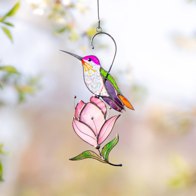 Stained glass hummingbird suncatcher with purple, green, and orange accents perched on a soft pink magnolia flower