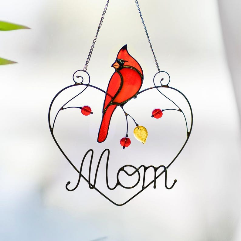 Stained glass cardinal suncatcher hanging on window with plant leaves, handmade bird decor glowing in sunlight, cozy home window hanging.