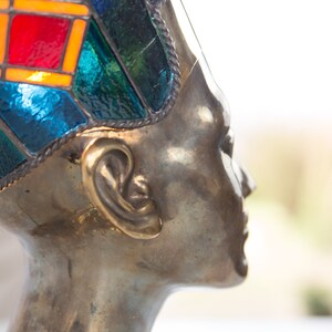 May include: A bronze sculpture of a person's head wearing a colorful stained glass crown. The crown is made of blue, green, red, and orange glass.