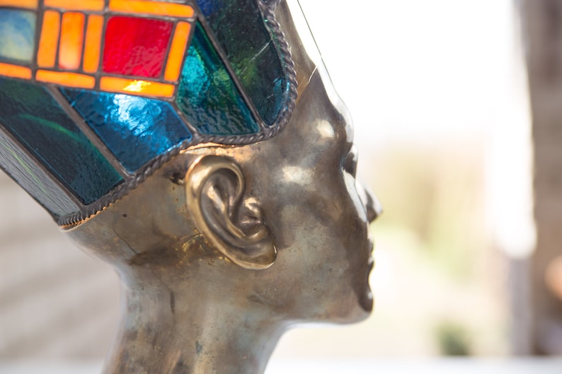 May include: A bronze sculpture of a person's head wearing a colorful stained glass crown. The crown is made of blue, green, red, and orange glass.