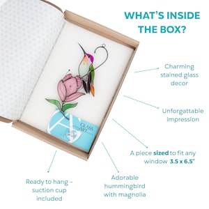Unboxing image of a vibrant stained glass hummingbird with a pink magnolia flower, nestled inside branded packaging with a suction cup and care card