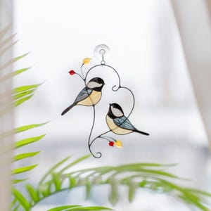 Stained glass bird suncatcher hanging on window with plant leaves, handmade chickadee glass decor catching sunlight, cozy window ornament.