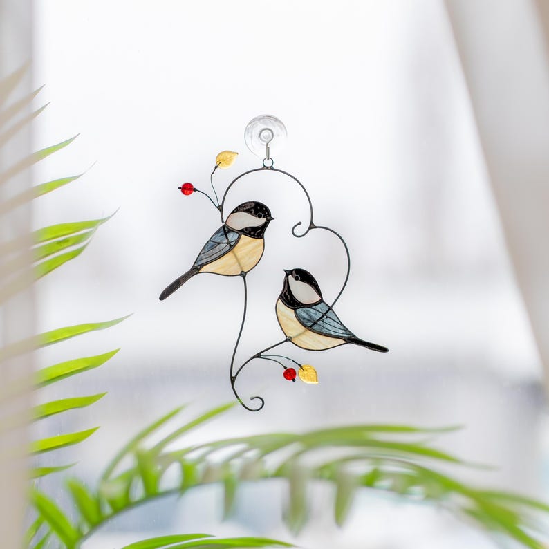 Stained glass bird suncatcher hanging on window with plant leaves, handmade chickadee glass decor catching sunlight, cozy window ornament.
