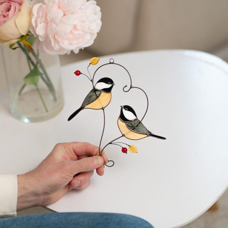 Stained glass chickadee couple suncatcher held by hand on table with flowers, handmade bird pair glass decor, love birds gift idea.