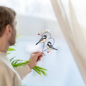 Stained glass chickadee pair suncatcher held by man near window, handmade bird couple window hanging with berries, romantic glass bird decor.