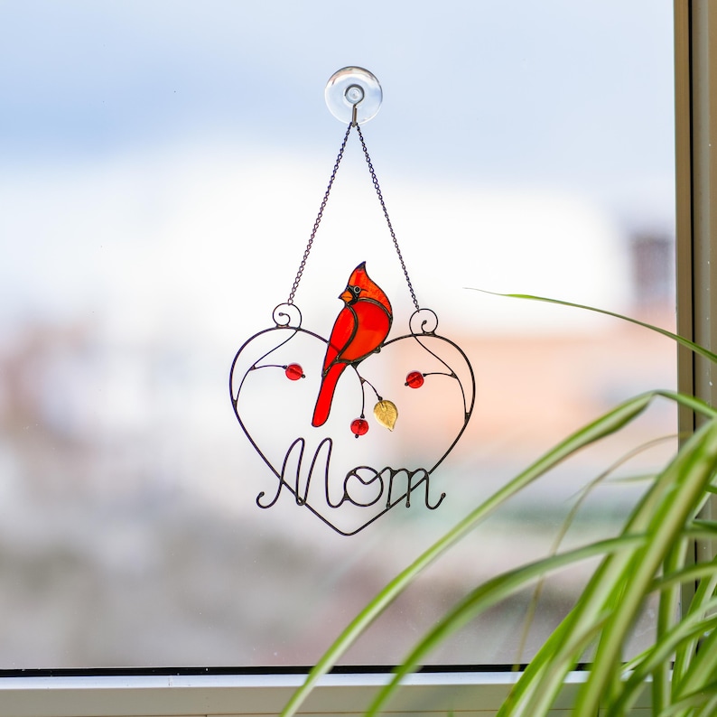 Stained glass cardinal suncatcher hanging on window with plant leaves, handmade bird decor glowing in sunlight, cozy home window hanging.
