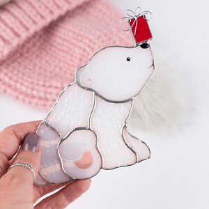 Hand holding a stained glass polar bear ornament balancing a red Christmas gift on its nose, with a soft pink knitted hat in the background.