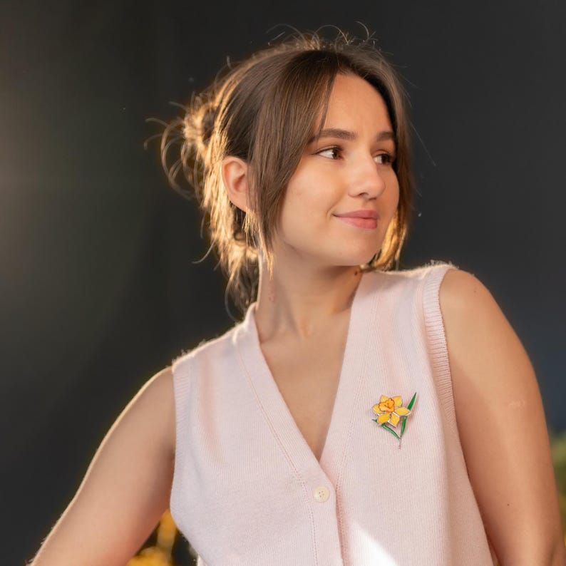 Woman wearing stained glass daffodil brooch in warm light, handmade yellow flower pin, delicate botanical jewelry styling.