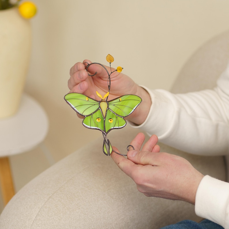 A detailed close-up of a person's hands holding a handmade Luna Moth suncatcher. This shot highlights the delicate wirework and the glowing lime-green stained glass textures.