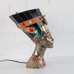 May include: A stained glass lamp shaped like the head of an Egyptian queen. The lamp has a black base with colorful geometric patterns and a gold trim. The queen's head is made of metal and has a detailed face.