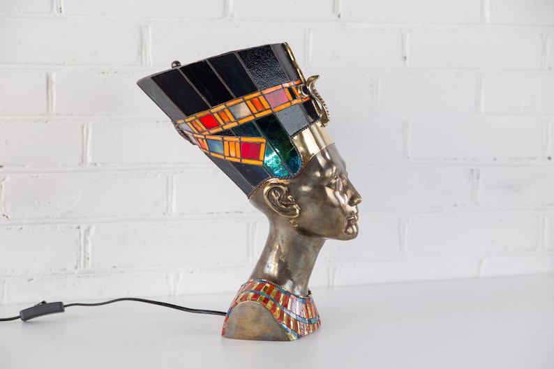 May include: A stained glass lamp shaped like the head of an Egyptian queen. The lamp has a black base with colorful geometric patterns and a gold trim. The queen's head is made of metal and has a detailed face.