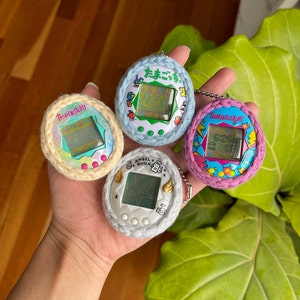 Puede incluir: Cuatro mascotas digitales Tamagotchi diferentes, cada una con un dise&ntilde;o y un color &uacute;nicos. Los Tamagotchi llevan todos cubiertas de ganchillo en diferentes colores, incluyendo rosa, azul, blanco y amarillo. Los Tamagotchi est&aacute;n todos en una mano, con un suelo de madera y una planta verde de fondo.