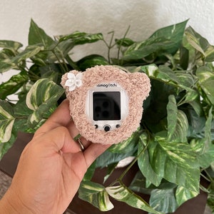 May include: A white tamagotchi with a brown fuzzy bear case. The case has a white flower on the top. The tamagotchi screen is black and white.