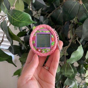 Puede incluir: Una funda de crochet rosa y verde para un juguete Tamagotchi morado. El Tamagotchi tiene una pantalla que muestra un laberinto y el texto "Tamagotchi".