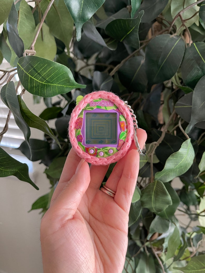 Puede incluir: Una funda de crochet rosa y verde para un juguete Tamagotchi morado. El Tamagotchi tiene una pantalla que muestra un laberinto y el texto "Tamagotchi".