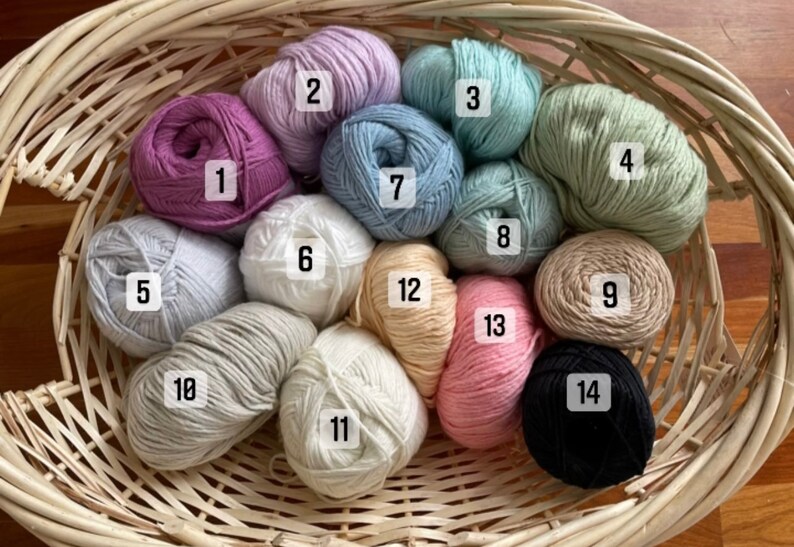 Puede incluir: Una cesta de mimbre llena de 14 ovillos de lana de varios colores, incluyendo azul claro, rosa, blanco, beige y verde. Cada ovillo est&aacute; numerado del 1 al 14.