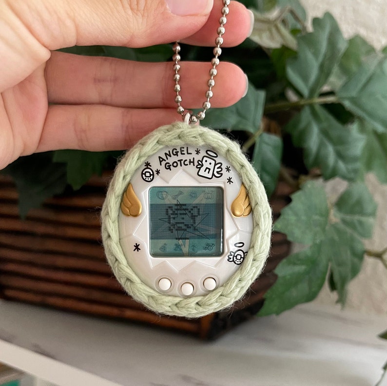 Puede incluir: Una mascota digital Angel Gotch blanca con una cubierta de crochet verde. La pantalla muestra una imagen pixelada. La mascota digital tiene una cadena dorada.