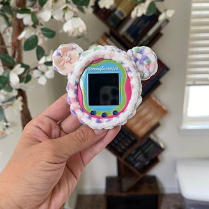 May include: A pink, blue and green Tamagotchi Uni toy with a fuzzy, white, pink and purple cover. The toy has a small screen and buttons.