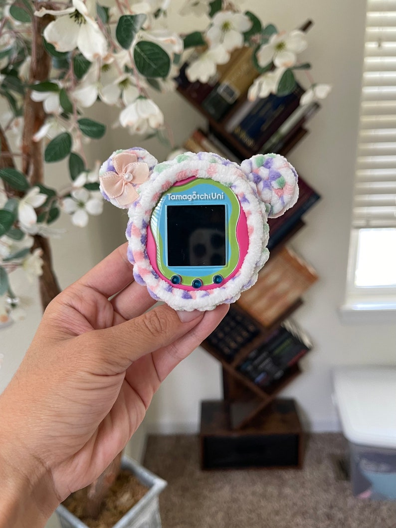 May include: A pink, blue and green Tamagotchi Uni toy with a fuzzy, white, pink and purple cover. The toy has a small screen and buttons.