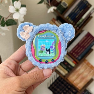 May include: A blue and pink Tamagotchi Uni toy with a fuzzy blue exterior and a pink screen. The screen displays a cartoon image of a dog and a house. The text "Tamagotchi Uni" is displayed on the screen.