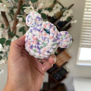 May include: A small crocheted plush toy in the shape of a bear's head. The toy is made with a white yarn with pink, purple, and green accents. The toy has a blue button eye.