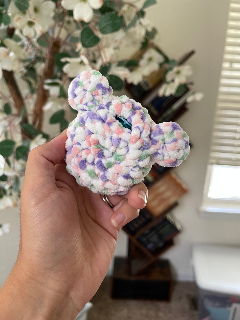 May include: A small crocheted plush toy in the shape of a bear's head. The toy is made with a white yarn with pink, purple, and green accents. The toy has a blue button eye.