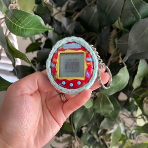 Puede incluir: Un juguete digital para mascotas Tamagotchi Connection rojo y amarillo con una cubierta de crochet azul y blanco. El juguete tiene una pantalla que muestra el texto "Tamagotchi Connection" y "6x16".