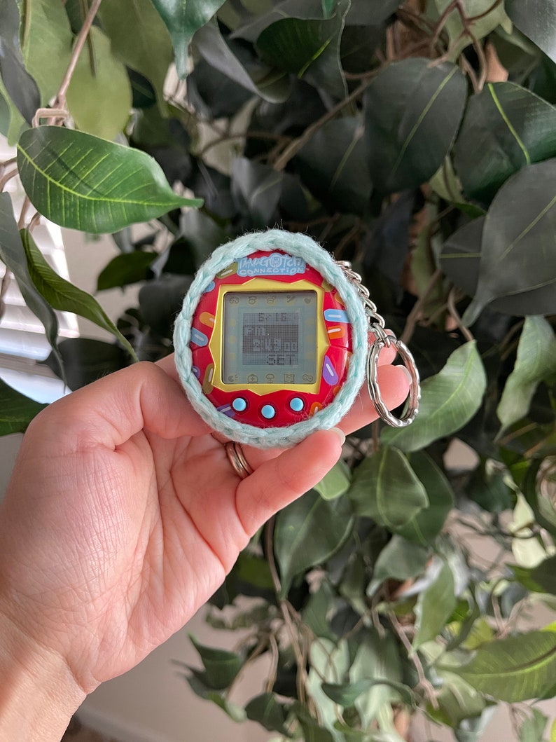 Puede incluir: Un juguete digital para mascotas Tamagotchi Connection rojo y amarillo con una cubierta de crochet azul y blanco. El juguete tiene una pantalla que muestra el texto "Tamagotchi Connection" y "6x16".
