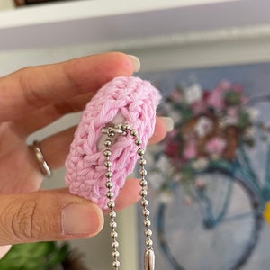 Puede incluir: Un llavero de crochet rosa con una cadena plateada y un cierre plateado. El llavero est&aacute; unido a un anillo plateado.