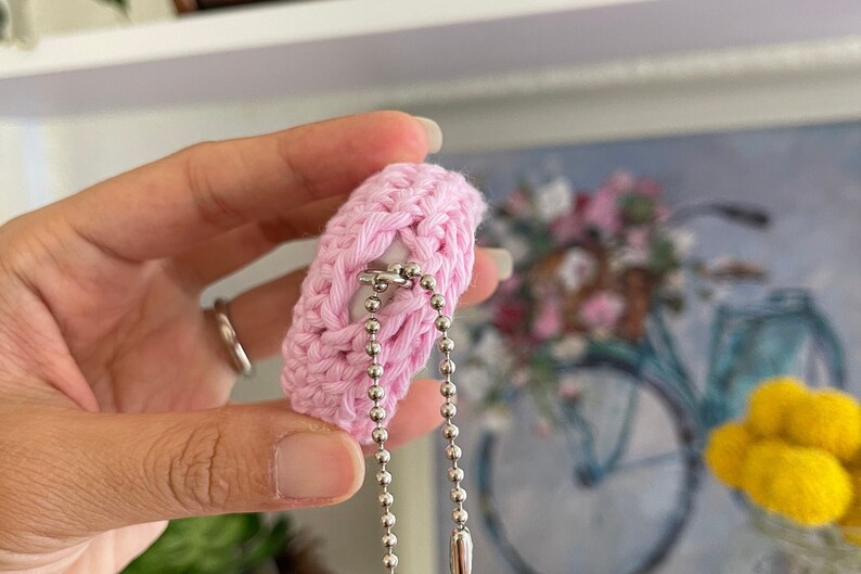 Puede incluir: Un llavero de crochet rosa con una cadena plateada y un cierre plateado. El llavero est&aacute; unido a un anillo plateado.