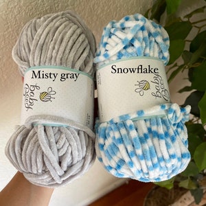 May include: Two skeins of yarn, one in a misty grey colour and the other in a white and blue snowflake pattern. The yarn is labelled "Baby Bee" and the colours are labelled "Misty Grey" and "Snowflake".