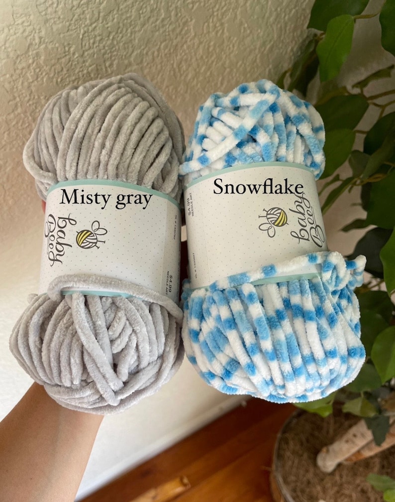 May include: Two skeins of yarn, one in a misty grey colour and the other in a white and blue snowflake pattern. The yarn is labelled "Baby Bee" and the colours are labelled "Misty Grey" and "Snowflake".
