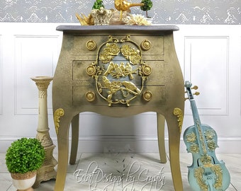 May include: A distressed gold and gray painted French style chest of drawers with three drawers. The chest has a decorative gold bird and floral design on the front. The chest is sitting on a tiled floor with a gold painted violin leaning against it.