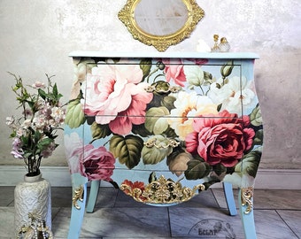 Puede incluir: Una cómoda de color azul claro con detalles dorados ornamentados y estampado floral. La cómoda tiene dos cajones y se asienta sobre cuatro patas. La cómoda está decorada con un patrón floral con rosas rosas, rojas y blancas. La cómoda está adornada con ribetes y herrajes dorados. La cómoda está sobre un suelo de baldosas.
