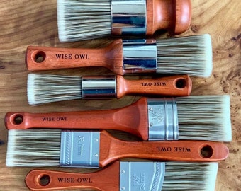 May include: Six paint brushes with wooden handles and silver metal ferrules. The brushes are arranged vertically, with the largest brush at the top and the smallest brush at the bottom. The brushes are all labeled "WISE OWL" and have different sizes, including 1/2", 3/4", 1", 1 1/2", and 2".