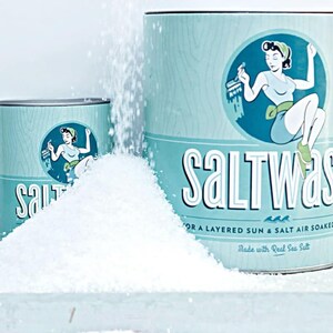 May include: A blue and white metal container with a vintage illustration of a woman holding a paintbrush. The container is labeled "Saltwash" and says "For a layered sun & salt air soaked look. Made with real sea salt."