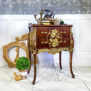 May include: A dark wood antique nightstand with gold accents and floral carvings. The nightstand has three drawers and ornate gold trim. An antique telephone and other decorative items sit on top. A gold picture frame and a small bird are in the foreground.