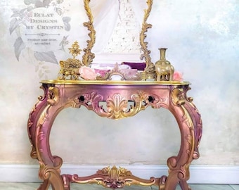 May include: A gold and pink ornate console table with a carved design. The table has a mirrored top and a gold ornate mirror above it. The table is decorated with a gold vase and a gold figurine.