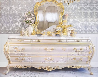 Könnte beinhalten: Eine aufwendig gestaltete Kommode mit Marmorplatte und goldenen Akzenten. Die cremefarbenen Schubladen sind mit floralen Mustern verziert. Ein goldgerahmter Spiegel und dekorative Elemente ergänzen das luxuriöse Design.