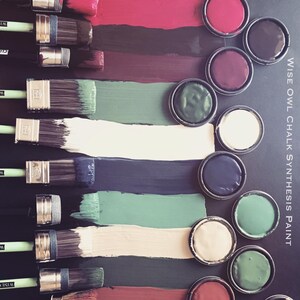 May include: A set of paint pots with various colors of chalk paint, including red, burgundy, green, black, white, and cream. The paint pots are arranged in a row on a black surface.  The paint is labeled "WISE OWL CHALK SYNTHESIS PAINT".