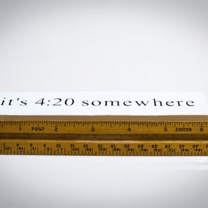 May include: A white wooden sign with black text that reads "it's 4:20 somewhere". The sign is sitting on top of a wooden ruler.
