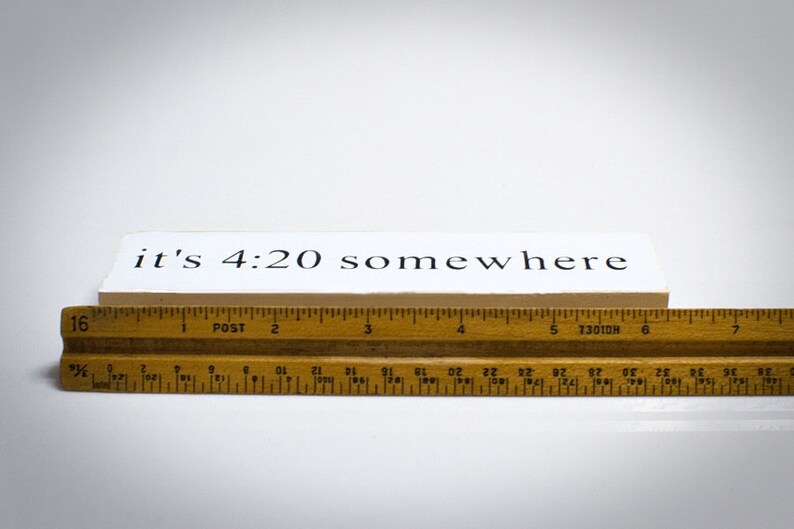 May include: A white wooden sign with black text that reads "it's 4:20 somewhere". The sign is sitting on top of a wooden ruler.