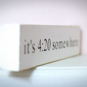 May include: White wooden sign with the text "it's 4:20 somewhere" in black lettering.