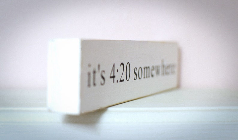 May include: White wooden sign with the text "it's 4:20 somewhere" in black lettering.