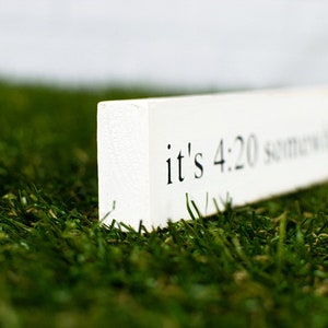 May include: A white wooden sign with the text "it's 4:20 somewhere" in black lettering, resting on a bed of green grass.
