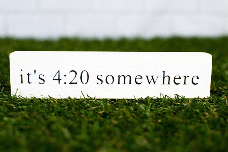 May include: White rectangular sign with black text that reads "it's 4:20 somewhere" sitting on green grass.
