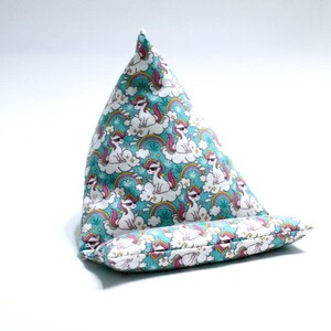 May include: A teal fabric book stand and a matching bolster pillow. The fabric features a repeating pattern of white unicorns wearing sunglasses, rainbows, and clouds. The book stand is triangular, and the bolster pillow is rectangular.